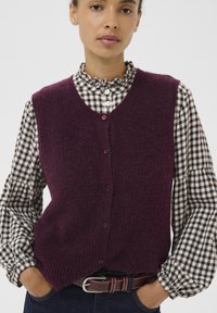 Bordeaux gebreide vest met een gestructureerde afwerking, voorzien van een V-hals en een sluiting met vijf knopen, gelegd over een zwart-wit geruit shirt.