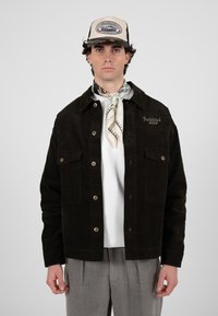 Junger Mann, der eine schwarze Cordjacke, ein weißes Hemd, graue Hosen, einen gemusterten Halstuch und eine beige Trucker-Cap vor einfachem Hintergrund trägt.