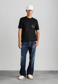 T-shirt preta com um bolso no peito apresentando riscas horizontais, combinada com jeans azul-escuro e sapatilhas brancas, finalizada com uma capa branca.