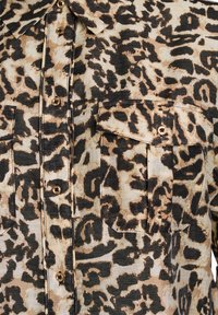 Chemise à souris léopard en tan et noir, avec deux poches avant et des boutons dorés. Tissu léger et texturé.
