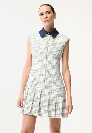 Kobieta ubrana w bezrękawnikową, białą sukienkę w niebieską kratę z ciemnoniebieskim kołnierzem i guzikami z przodu, stojąca na tle jednolitego tła.