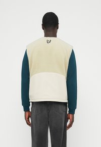 Gilet in pile color crema e beige con superficie testurizzata. Indossa una camicia a maniche lunghe blu sotto e presenta un logo ricamato sulla schiena.