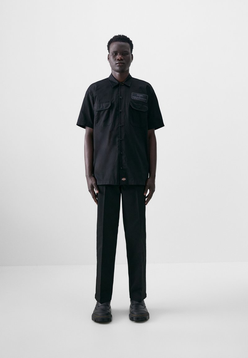 Zwarte shirt met korte mouwen, twee voorkruiszakken, knoopsluiting; gecombineerd met zwarte rechtgesneden broek en zwarte schoenen, gladde stoftextuur.