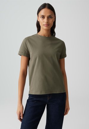 Vrouw met lang donker haar, gekleed in een olijfgroen T-shirt met korte mouwen en een donkere spijkerbroek, staand tegen een effen grijze achtergrond.