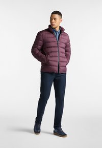 Chaqueta de plumas burdeos con detalles de costura horizontal, cierre de cremallera y cuello, combinada con camisa azul y pantalones azul marino, completada con zapatillas.
