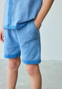 Lichtblauwe gebreide outfit met een shirt met korte mouwen en knopen, en een gebloemd short met omgeslagen mouwen en zijzakken.