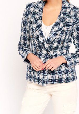 Vrouw draagt een marineblauw-wit geruit double-breasted blazer met metalen knopen, een wit topje en crèmekleurige broek, handen in elkaar gevouwen ter hoogte van de taille.