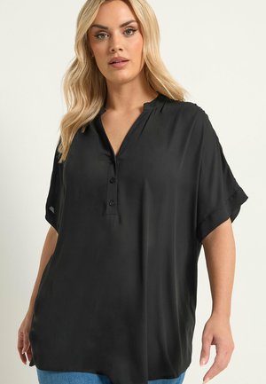 Lyshåret kvinde iført løs sort kortærmet bluse med knappet V-udskæring og blå jeans, stående mod en ensfarvet lys baggrund.