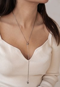 Zilveren hangerketting met een druppelontwerp, uitgerust met een dunne ketting en een ronde kraalaccent, gedragen over een licht geribbelde trui.