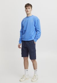 Mėlynas džemperis su priekiu kišene, derinamas su tamsiai mėlynomis kroso stiliaus šortais. Aksesuarai: balti sportbačiai ir kojinės iki blauzdos. Paprastas dizainas.