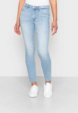 Person, der hellblaue Slim-Fit-Jeans und weiße Turnschuhe trägt, vor einem schlichten hellen Hintergrund steht.