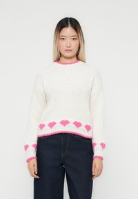 Jauna sieviete valkā baltu adītu džemperi ar rozā sirdīm ap apakšmalu un piedurknēm, kopā ar tumši ziliem džinsiem, stāvot pret baltu fona.