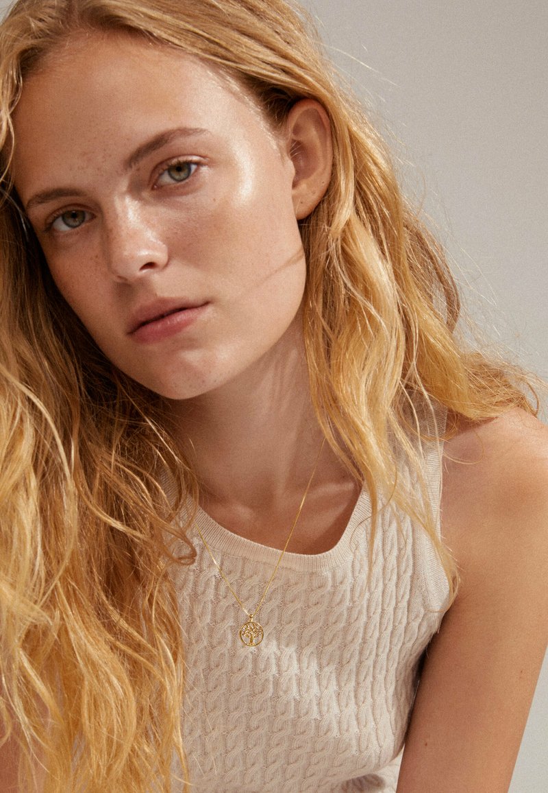 Top in maglia di un beige chiaro con un motivo testurizzato, impreziosito da una collana dorata con un pendente a forma di albero; i capelli biondi e mossi incorniciano il viso.