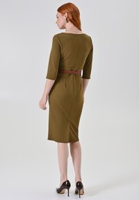 Olijfgroene aangesloten jurk met driekwart mouwen, een getailleerde taille met ceintuur en een split aan de achterkant, gemaakt van gladde stof met een subtiele glans.