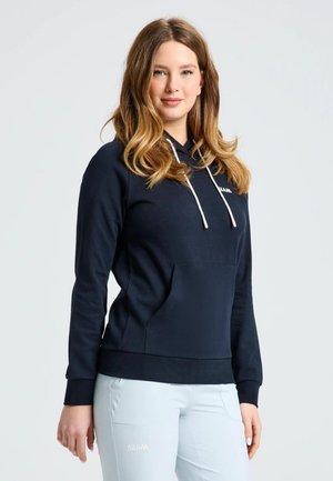 Una mujer posa con una sudadera azul marino con capucha y cordones blancos, y unos pantalones claros, sobre un fondo liso y claro.