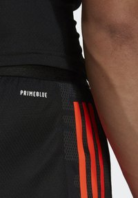 Svarta träningsshorts i strukturerad tyg, med röda vertikala ränder och en "PRIMEBLUE"-logotyp. Närbild på sida söm.