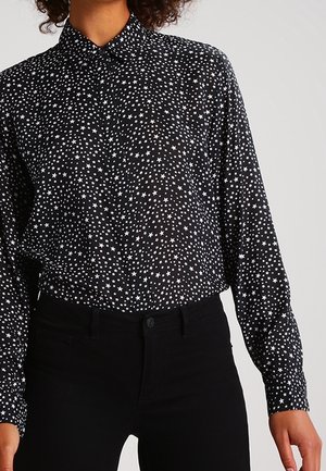 Musta napillinen paita, jossa valkoiset tähtikuviot, mustien housujen kanssa, käytetty henkilöllä yksiväristä taustaa vasten.