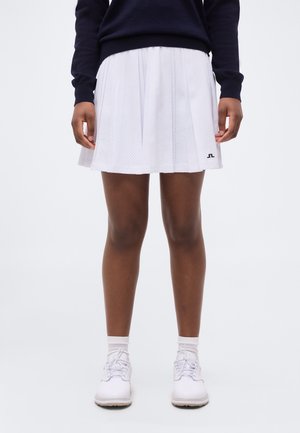 Άτομο που φορά λευκή πιετέ φούστα, λευκά αθλητικά παπούτσια και μπλε μακρυμάνικη μπλούζα, όρθιο μπροστά σε απλό φόντο.