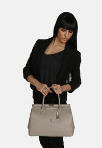 Beżowa skórzana torebka o usztywnionym kształcie, z podwójnymi uchwytami na górze i metalowymi akcentami. Zauważalny detal z metką zawieszony jest na jednym z uchwytów.