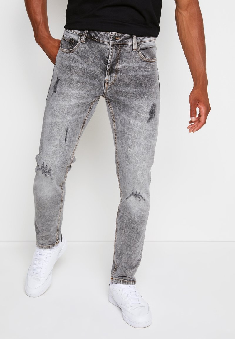 Homme portant un jean gris délavé et usé, des baskets blanches et une chemise noire, debout devant un fond blanc uni.
