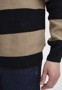 Strikkegenser med brede striper i beige og svart, ribbestrikkede mansjetter og kant. Materialet ser teksturert ut med en løs passform.