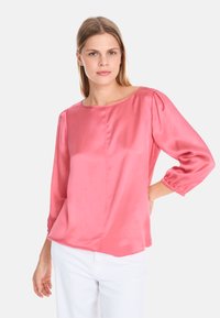 Rosa Satinbluse mit Dreiviertelärmeln, Rundhalsausschnitt und gerafften Manschetten. Trägt man mit weißen Hosen, die eine glatte, glänzende Textur zeigen.