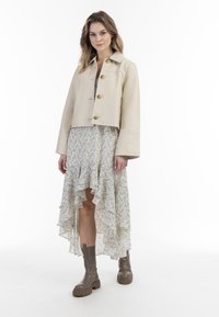 Beige lässige Lederjacke mit zwei goldenen Knöpfen, kombiniert mit einem cremefarbenen, rüschengesäumten Maxikleid mit floralem Muster und kniehohen braunen Stiefeln.