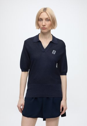 Femme blonde aux cheveux courts portant un polo tricoté bleu marine à manches courtes et un short bleu marine assorti sur fond blanc.