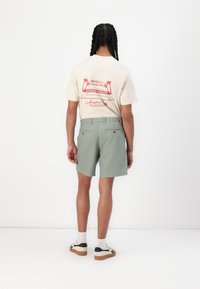 T-shirt beige clair avec impression graphique rouge ; shorts verts, devant plat, deux poches au dos, associés à des chaussures blanches et des chaussettes de cheville.