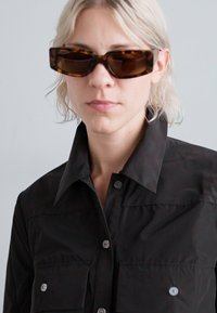 Black button-up shirt with a collar, featuring two front pockets, made of smooth fabric. Model wearing brown tortoiseshell sunglasses.