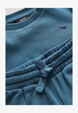 Blå bomuldssweatshirt med ribstrikket hals; matchende sweatpants med elastik i taljen og snøre. Broderet logo i mørkere blå.