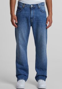 Jeans de mezclilla azul con corte relajado, que cuentan con un cierre de botón, cinco bolsillos y ligero desgastado. Combinados con zapatillas deportivas blancas.