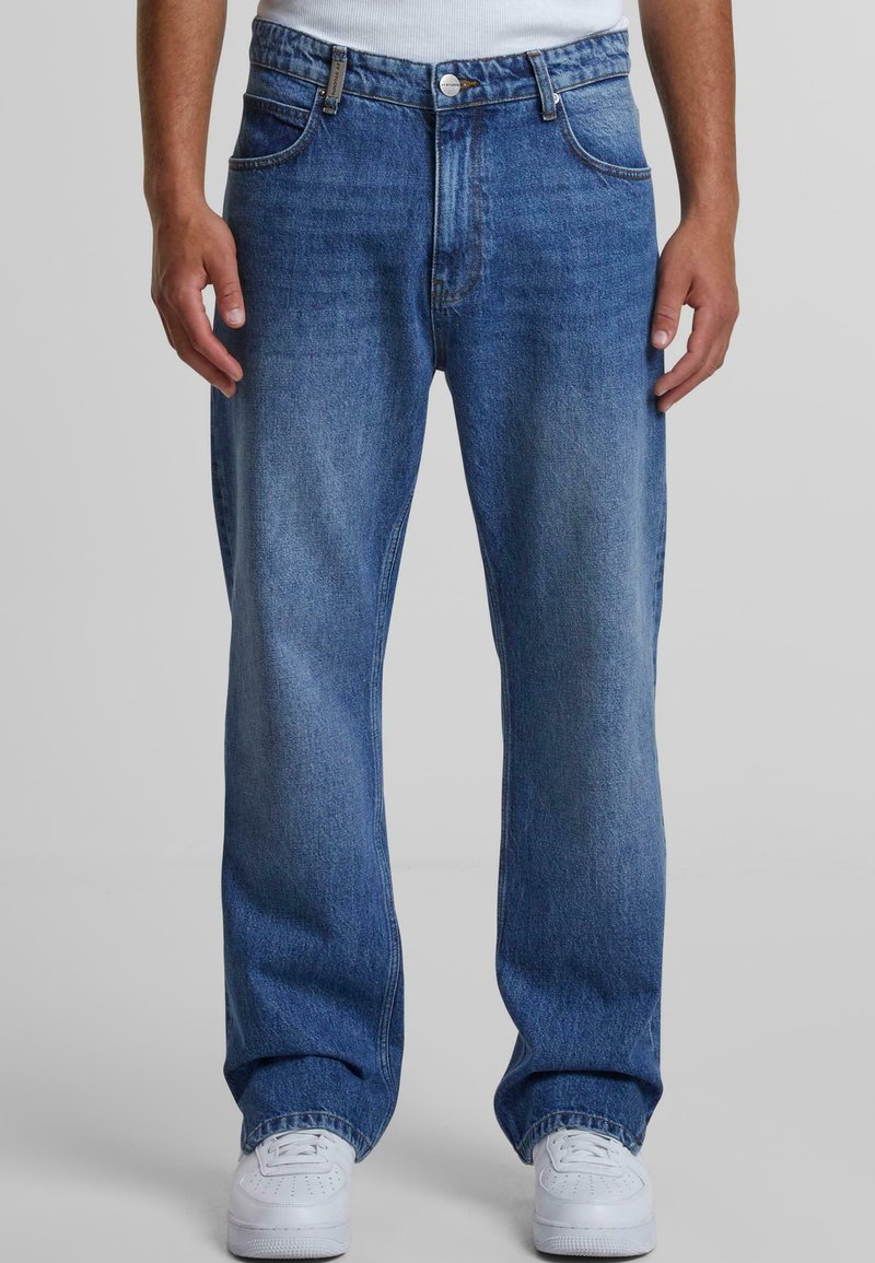 Jeans in denim blu con vestibilità rilassata, dotati di chiusura a bottoni, cinque tasche e leggera sbiaditura. Abbinati a scarpe sportive bianche.
