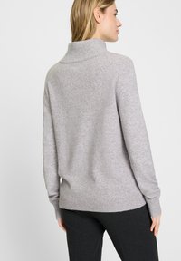Grijze gebreide trui met een hoge kraag, een losse pasvorm, geribbelde manchetten en zoom. Zachte textuur, gecombineerd met donkere broek, gezien vanuit de achterkant.