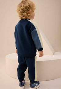 Granatowy dres z jasnoniebieskimi i kremowymi akcentami. Bluza ma rękawy raglanowe; spodnie mają boczne pasy. Zestaw zawiera szare sneakersy.