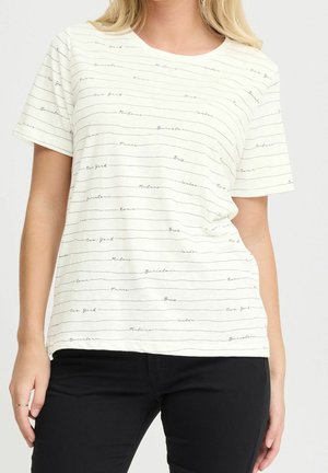Vrouw die een wit T-shirt draagt met dunne zwarte horizontale strepen en stadnamen zoals New York, Parijs en Berlijn, gecombineerd met een zwarte broek.