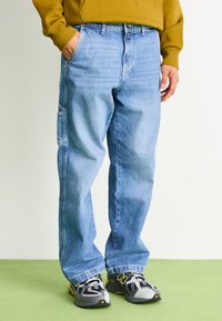 Light blue denim jeans with a relaxed fit, featuring a side pocket and subtle fading, paired with dark athletic shoes.