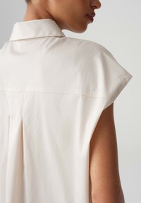 Lichtbeige shirt met een gespreide kraag, mouwloze cap sleeves en een plooidetail aan de achterkant, die de soepele stoftextuur en minimalistisch ontwerp benadrukt.