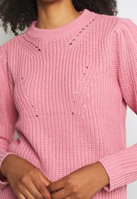 Pull rose en tricot présentant une texture côtelée, un col rond et de petites découpes décoratives le long de l'encolure et des côtés.