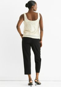 Woman standing with hands on hips wearing a sleeveless white sequined top, black cropped pants, and black heeled sandals on white background.