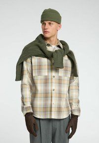 Béžová a hnědá kostkovaná košile, olivově zelená pletená čepice, odpovídající šála, hnědé rukavice, šedé kalhoty. Neformální zimní outfit.