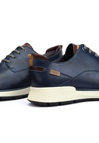Chaussures en cuir bleu marine avec un design perforé, des lacets gris et des accents marron. Semelle blanche épaisse avec texture nervurée. Logo sur le côté.