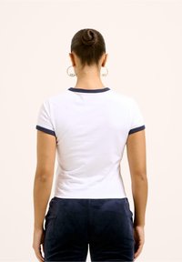 Vit kroppsnära t-shirt med marinblå kant runt kragen och ärmarna, med en slät textur och enkel design. Mörka byxor synliga.