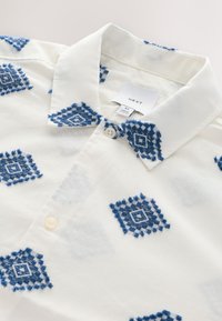 White collared shirt with blue embroidered diamond patterns and cream buttons, labeled medium size from NEXT brand.