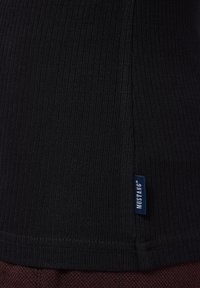 Schwarzer, gerippter Stoff mit strukturierter Oberfläche und einem seitlichen blauen Etikett, das ein kleines Markenlogo zeigt. Der untere Saum ist mit einer Naht versehen.