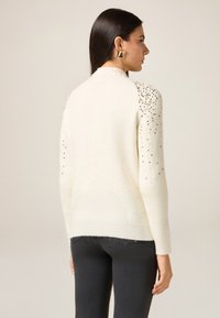 Samtweicher Pullover in Creme mit Rippstrickstruktur und Paillettenverzierung auf den Schultern. Ausgestattet mit langen Ärmeln und einer lockeren Passform.