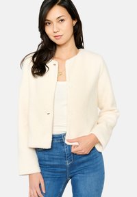 Crèmekleurige, gestructureerde jas met een ronde halslijn, knoopsluiting en lange mouwen, gecombineerd met een aansluitend wit topje en blauwe jeans.