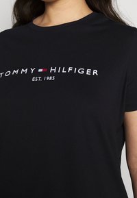 Camiseta de manga corta negra con bordado blanco que dice "TOMMY HILFIGER EST. 1985" y pequeño logo de bandera roja, blanca y azul en el pecho.