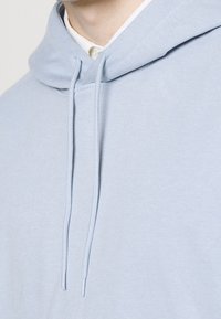 Sudadera azul claro hecha de tejido suave, que presenta un cordón ajustable, un bolsillo canguro estándar y un corte holgado con una textura suave.