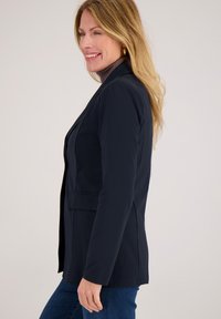 Marineblauw colbert van gladde stof, met een getailleerde snit, een knoopsluiting en twee voorzakken. Gestyled met een coltrui.